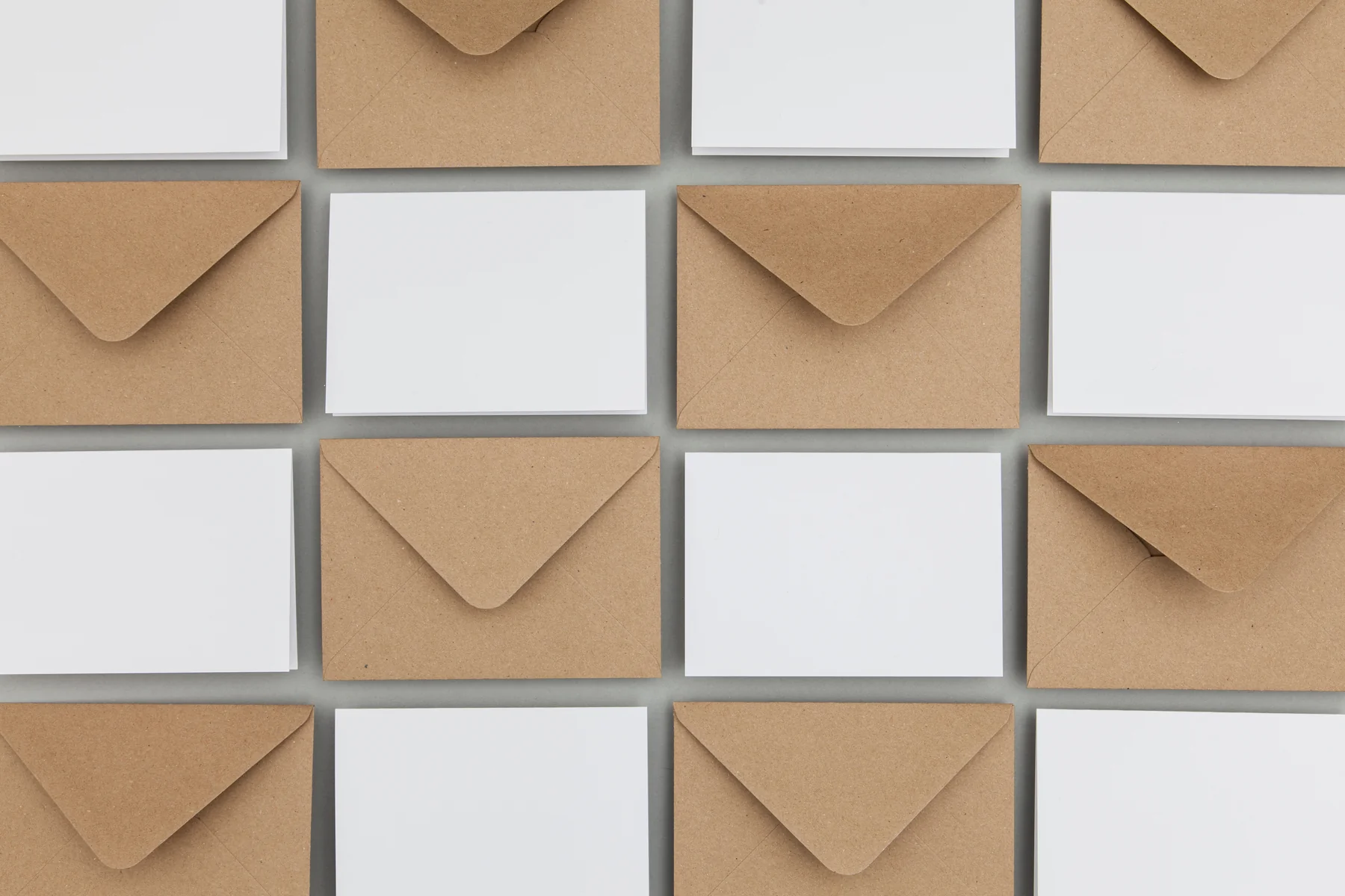 Flat-lay grid pattern of blank envelopes alternating between brown kraft paper and white, arranged diagonally on a gray surface.